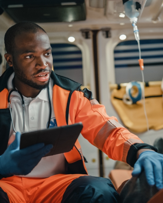 a medical professional sits in the back of an ambulance and uses a tablet while talking to an off camera patient